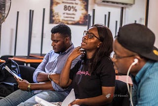 Oladoyin Idowu is sitting next to two men and she is wearing glasses. Her mouth is open as she is explaining a concept.