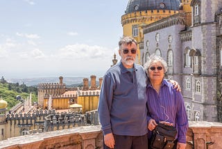 The author and his wife on a balcony of the Pena Palace in Sintra, Portugal with a view in the background of a large portion of the palace.