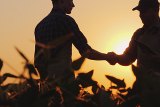 Two farmers stand in a field and shake hands in silhouette at sunset.