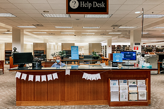 The help desk at the BYU Family History Library — October 15, 2020. Long desk holding computer monitors atop it, and a “WELCOME BACK” banner hanging underneath the top of it, with a male masked staffer sitting behind it.