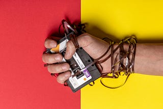 Photo of a hand clutching an audiocassette tape, with the tape dragged out from the cartridge and wrapped around the hand and wrist, against a pink and yellow background.