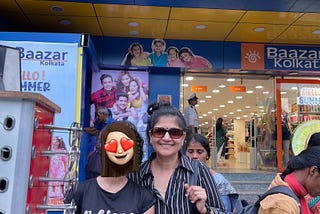 A woman with brand new sunglasses shows them off. A girl, around 16, is next to her. Her daughter! The girl’s face is covered with an emoji sticker. A signboard says, “Bazaar Kolkata” behind them.