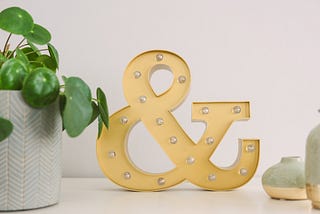A large ampersand sits on a clean desk between a potted plant and a vase.