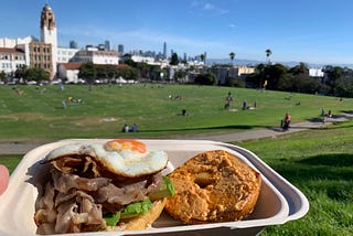 A hand holding a to-go container with the aforementioned egg sandwich in front of an open field at the park.