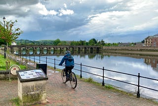 A person riding a bike along the side of a river with a bridge in the background.