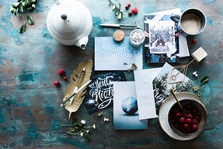 An aerial view of a writing desk, with a pen, a feather, several holiday cards, a teapot and cup of chocolate and a bowl of berries all arranged together.