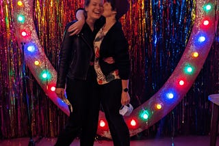 Two women with dark hair are embracing, one is kissing the other on the cheek and they both look joyful. They stand in front of a large heart illuminated with colourful lights