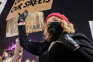Several women protesters standing in the street, one blonde woman with a black mask holding a sign saying, “Our Bodies, Our Streets.”