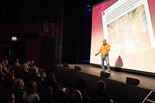 Gordon Fong standing on stage with a microphone in hand explaining about the tweet behind him on a big screen.