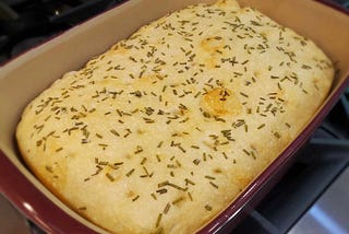 Image of baked focaccia, topped with sea salt and fresh rosemary, in stoneware pan