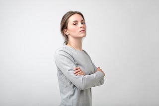 Young woman standing looking confident or arrogant in front of gray background, crossing her arms across her chest.