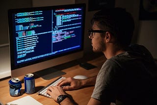 Software Developer sitting in a dimly lit room in front of the computer typing on keyboard and looking at code.