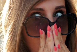 Blonde woman has a surprised face. She is looking over her big sunglasses and she is holding both hands (with painted fingernails) in front of her face.