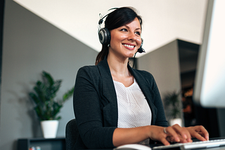 A smiling woman with a headset types at her computer.