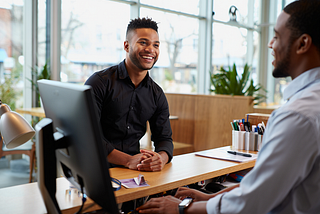 Two colleagues at a desk — one smiling and making eye contact with a customer while the other types into a computer system.