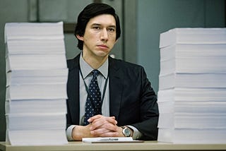 Still from the movie ‘The Report’ shows Dan Jones, played by Adam Driver, a Senatorial investigator at his desk in front of 2 towers of paper.