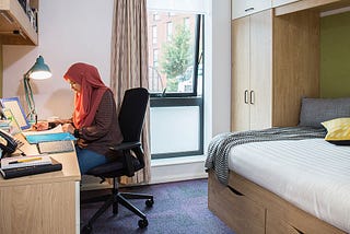 a female student in university accommodation. she is sitting at a desk and writing next to a lamp.