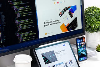 A white desk with a large monitor, ipad, iphone and Starbucks coffee cup. The large monitor displays a text editor with code next to a landing page of a website.