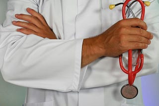 A man’s crossed arms; the white coat and stethoscope suggest it’s a doctor