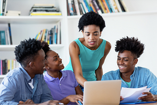 three students working together on one laptop while a teacher overlooks.