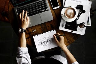 Person with computer, a sketchpad with words “Pass it on”, latte, iPhone, earphones, and magazines