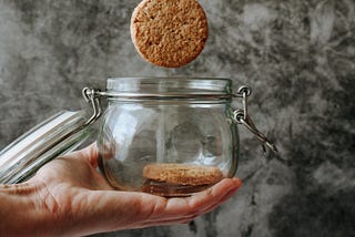 Hand holding cookies jar with one cookie inside and one falling into the jar.