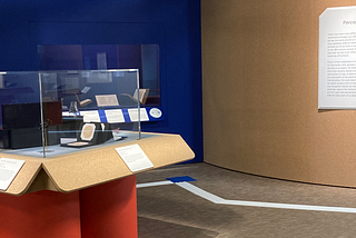 A photograph of the Wellcome Collection “In plain sight” exhibition featuring a glass cabinet on a red stand. The floor shows a brown fabric flooring with tactile tape used to guide those with visual impairments around the installation. The walls are textured with cord board to provide physical feedback for people with visual imparement.
