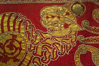 Detail of an embroidered mantle. The fabris is dyed deep red and gold embroidery depicts a stylised lion with a mane that looks like flames and a striped body. The finer details are picked out in double rows of pearls.