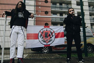 Bob Vylan lean against a construction fence, a St. George’s flag with their logo on it between them.