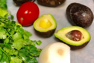 Ingredients to make guacamole. Cilantro, cut onion, avocado split in half, roma tomato on a plastic cutting board.