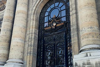 Color photograph of the MAH’s monumental entrance with large stone columns. Above the ornate black wrought-iron door, a complex decoration with gilded sculptures including a central figure. Engraved names at top: ‘TOEPFFER’ and ‘LIOTARD