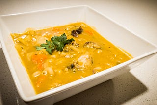 A white square bowl showing soup joumou with chicken legs, carrots, and pasta, garnished with fresh parsley in the center.