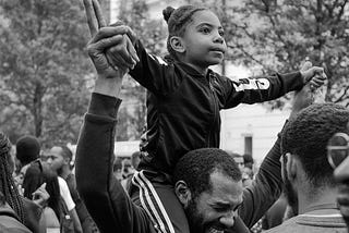 A young Black girl sits on a Black man’s shoulders (presumably her father), extending her arms as she holds his hands and looking hopeful among a crowd of Black protestors.