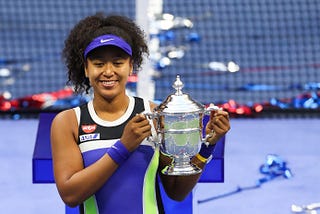 Head to mid body shot of Naomi Osaka, wearing bright blue and green athletic clothing, holding a trophy from a tennis tournament, and smiling.