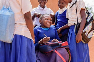 A young girl in a blue sweater sits in a wheelchair smiling surrounded by other school friends in white and blue uniforms all smiling