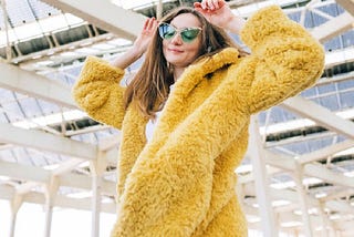 A woman wearing a yellow fake fur coat, green lensed sunglasses, and feeling herself.