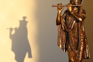 A full length Krishna statue is placed to the right of the image with sunlight falling on it in an angle from the top making it shine and there is a shadow of the statue falling to the left of the image.