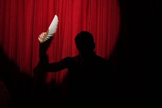 A silhouette with a feather in their hand. In the background there is a red curtain.