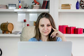 A smiling woman sitting in front of a bookshelf, looking down at her laptop with one finger resting on her face.