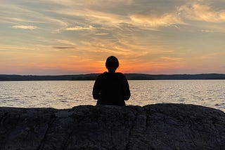 The author is sitting by a lake, facing the sunset in Sweden