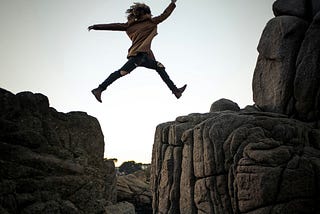 A person jumping from a large boulder to another large boulder over a sudden drop.