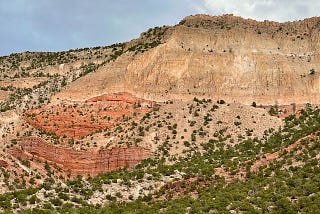This is a landscape on the Jemez Pueblo in New Mexico. The grey brown sandstone of the mesa contrasts with a light blue sky and clouds. Beneath this grey brown sandstone is Navajo red sandstone, then below that is dotted with juniper, cholla cacti and other desert vegetation.