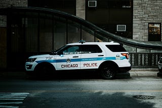 A Chicago Police SUV parked along a city street at night outside a stone and glass building, its blue-and-white markings and “Chicago Police” lettering clearly visible under dim street lighting.