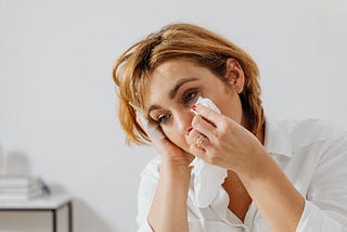 Woman with medium length dark blond hair wearing a white button down shirt. She is dabbing her eye with a tissue because she has been crying.