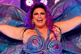 A screenshot of a smiling Spankie Jackzon with her arms outstretched from season 2 of RuPaul’s Drag Race Down Under, wearing a purple, blue and pink handcrafted costume that somewhat resembles a butterfly.