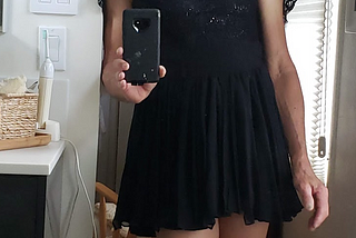 Picture of Miri standing in a doorway, wearing a very cute short black dress, smiling and holding a camera to take the picture of herself. She is hoping you like what you see.