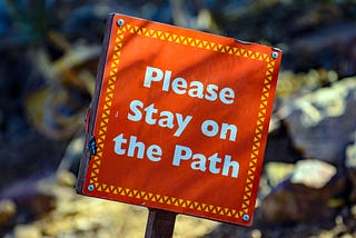 A picture of a red sign on a stake driven into the ground that says “Please stay on the path”.