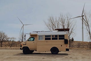 A small tan bus which has been converted into a tiny home, or skoolie, is parked in front of two large wind turbines.