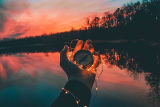 A compass surrounded by fairy lights and held by someone whose arm and palm can be seen, while behind it is a lake and a beautiful sunset.
