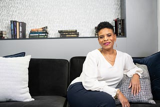 Picture of Stacey Mahoney sitting on a sofa smiling. She is wearing a white shirt and navy pants. Medium brown skin with short cropped hair.